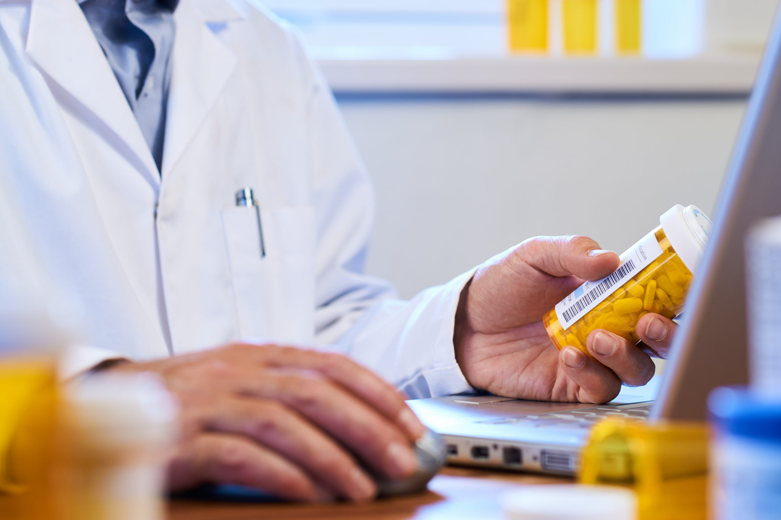 Person im Arztkittel am Laptop, eine Hand auf der Computermaus, in der anderen Hand eine Dose mit Tabletten